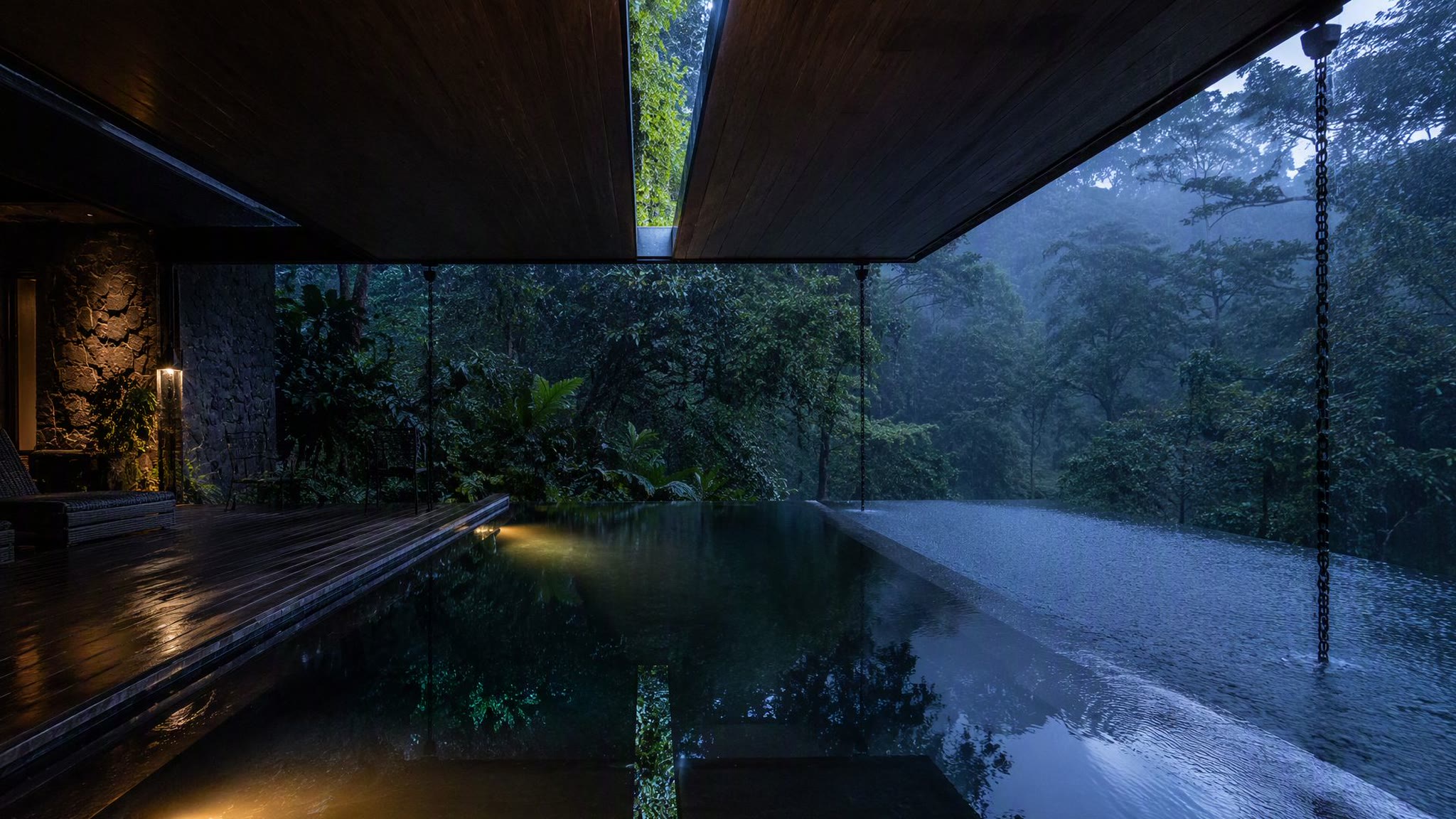Plunge pool half under roof, half under sky, rain dimpling the surface at blue hour.