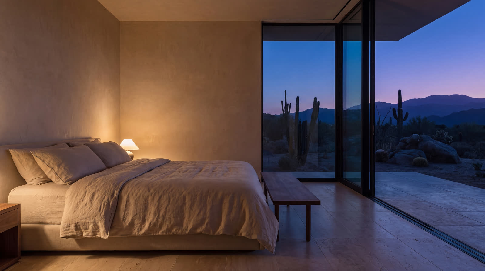 A low platform bed in linen against plaster, a glazed wall open to violet dusk, saguaro and mountains beyond.