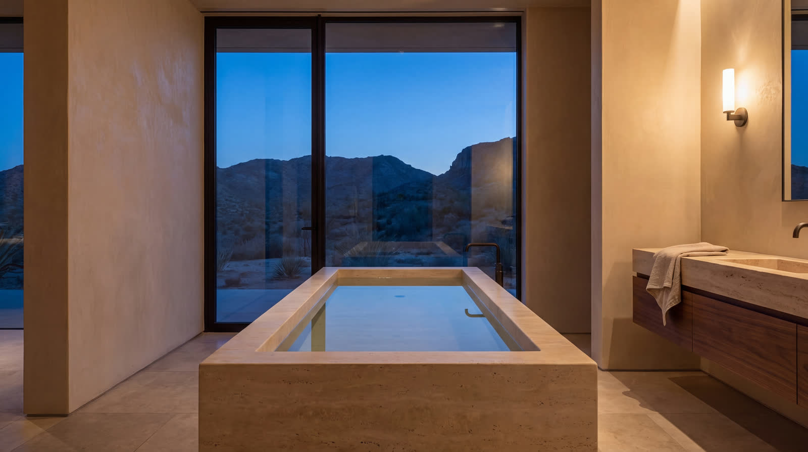 A monolithic travertine soaking tub before a full-height window onto darkening desert, walnut vanity beside it.